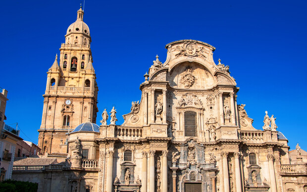 A grand baroque cathedral features ornate sculptures and carvings on its facade, with a towering bell tower rising beside it. The surroundings are clear under a bright blue sky.